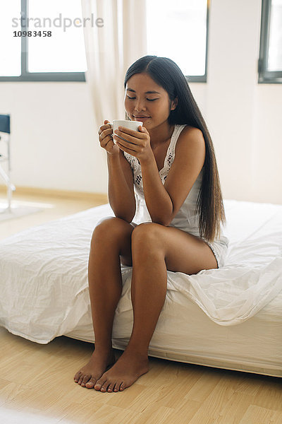 Junge Frau sitzt auf dem Bett und trinkt Kaffee.