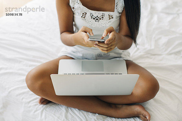 Junge Frau auf dem Bett sitzend mit Handy und Laptop