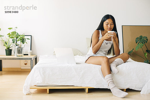Junge Frau sitzt auf dem Bett und trinkt Kaffee.