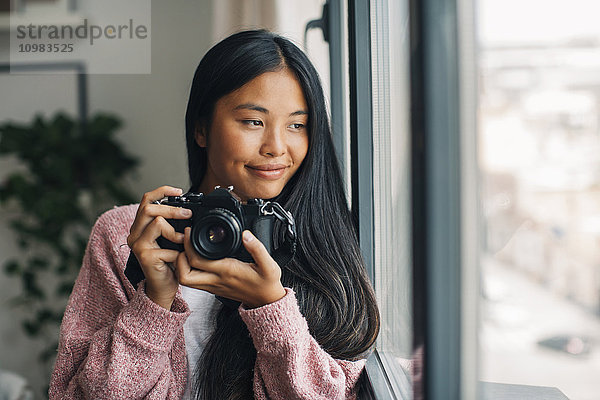 Porträt einer lächelnden jungen Frau mit Kamera durchs Fenster schauend