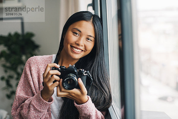 Porträt einer lächelnden jungen Frau mit Kamera in Fensternähe