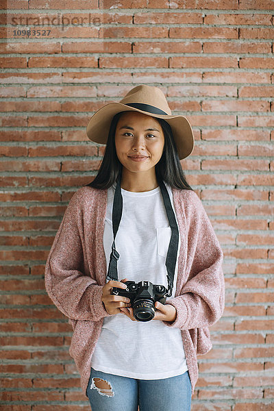 Porträt der lächelnden jungen Frau mit Kamera vor der Ziegelwand
