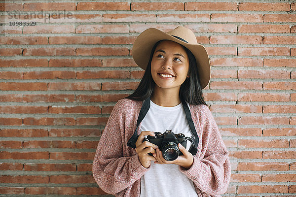 Porträt der lächelnden jungen Frau mit Kamera vor der Ziegelwand