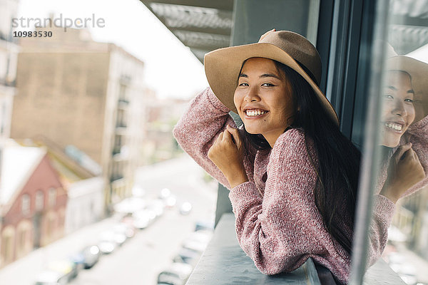 Porträt einer lächelnden jungen Frau mit aus dem Fenster gelehntem Hut
