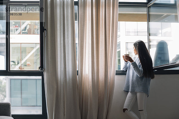 Junge Frau mit einer Tasse Kaffee durchs offene Fenster schauend