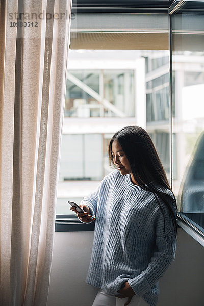 Junge Frau mit Tasse Kaffee vor offenem Fenster mit Blick aufs Handy