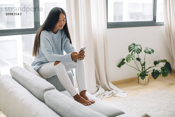 Junge Frau sitzt auf der Couch im Wohnzimmer mit Smartphone