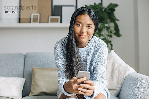 Porträt einer lächelnden jungen Frau mit Smartphone auf der Couch zu Hause