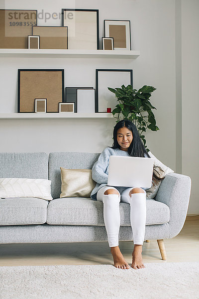 Lächelnde junge Frau sitzt auf der Couch zu Hause mit Laptop