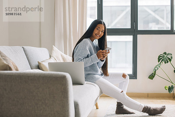 Lächelnde junge Frau sitzt auf der Couch mit Smartphone