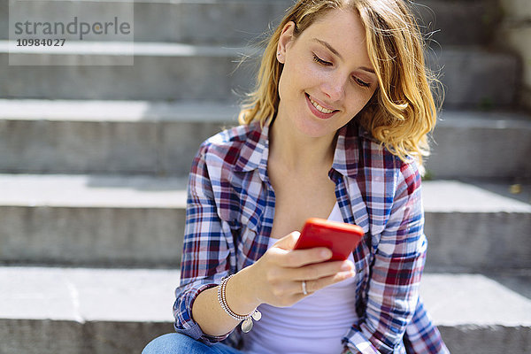 Porträt einer lächelnden jungen Frau  die mit dem Smartphone auf der Treppe sitzt.