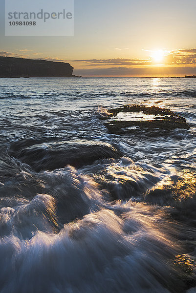 Australien  New South Wales  Seelandschaft mit rauschendem Wasser und aufgehender Sonne und Gegenlicht  Wellen