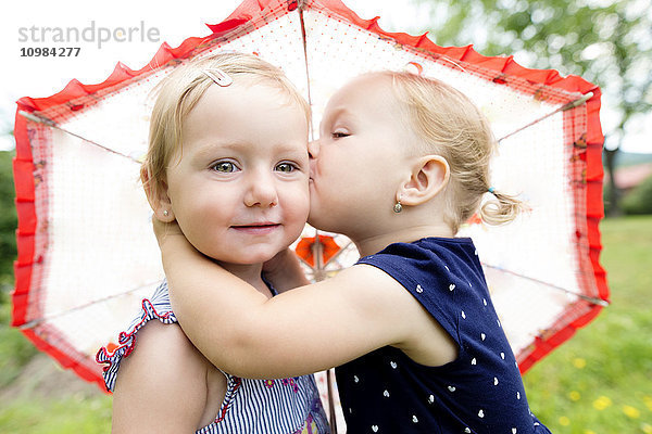 Kleines Mädchen  das seine Schwester unter einem Regenschirm küsst.