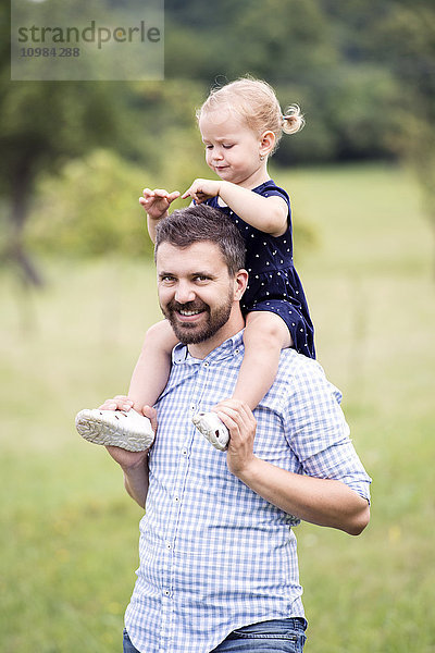 Glücklicher Vater trägt kleine Tochter auf den Schultern in der Natur