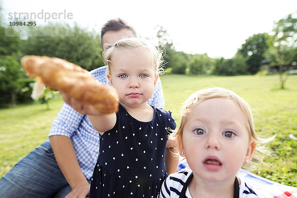 Kleines Mädchen mit Vater und Schwester in einem Park  das dem Betrachter ein Croissant anbietet.