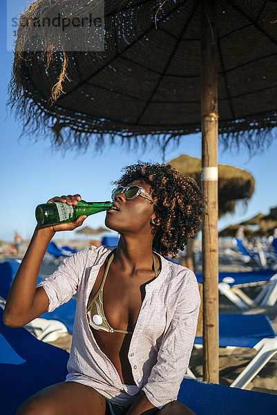 Junge Frau sitzt am Strand und trinkt Bier.