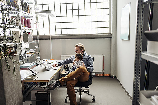 Mann mit Baby auf dem Schoß mit Laptop am Schreibtisch in einer Fabrik