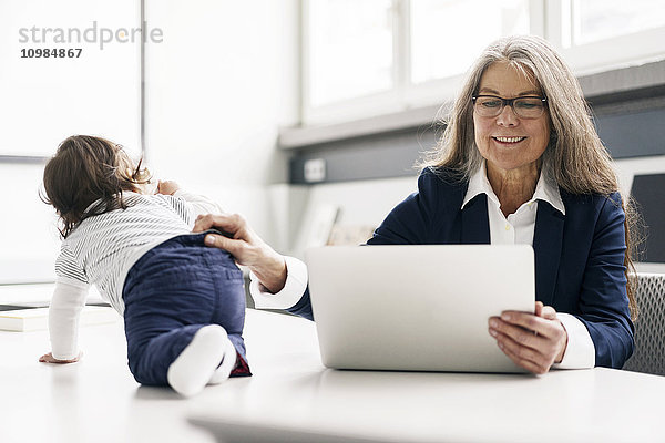 Senior Geschäftsfrau sitzend am Konferenztisch mit Laptop und Baby Girl