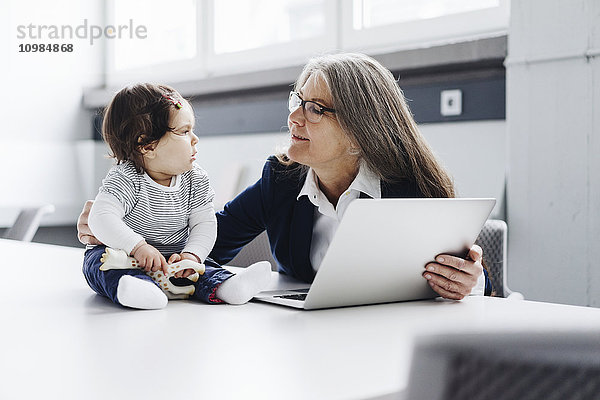 Senior Geschäftsfrau sitzend am Konferenztisch mit Laptop und Blick auf Baby Girl