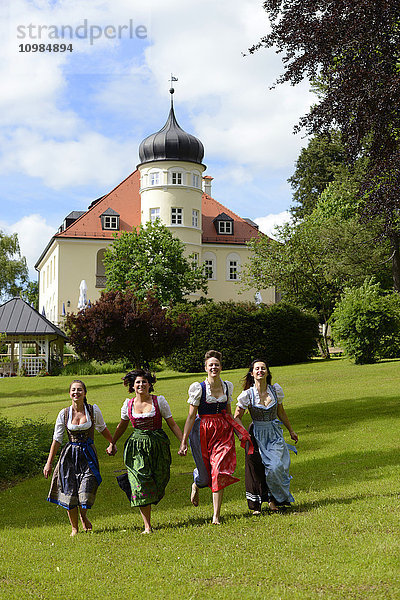 Deutschland  Bad Heilbrunn  vier glückliche Frauen mit Dirndln  die Hand in Hand auf einer Wiese laufen.