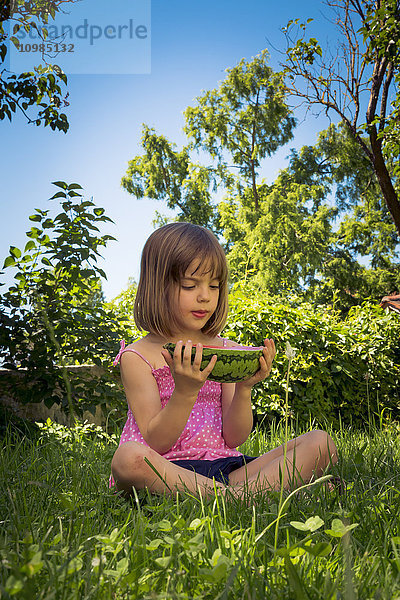 Kleines Mädchen sitzt auf einer Wiese und hält eine Scheibe Wassermelone.
