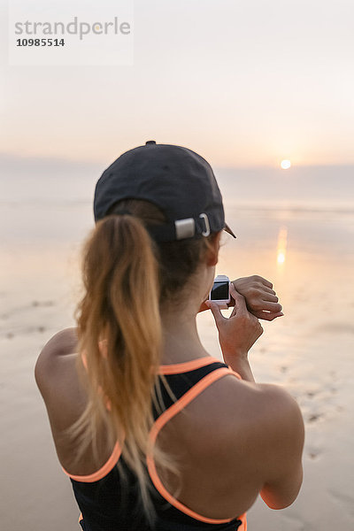 Junge Sportlerin mit Blick auf die Smartwatch am Strand bei Sonnenuntergang
