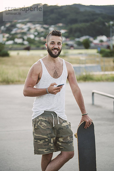 Porträt eines lächelnden Mannes mit Skateboard und Smartphone