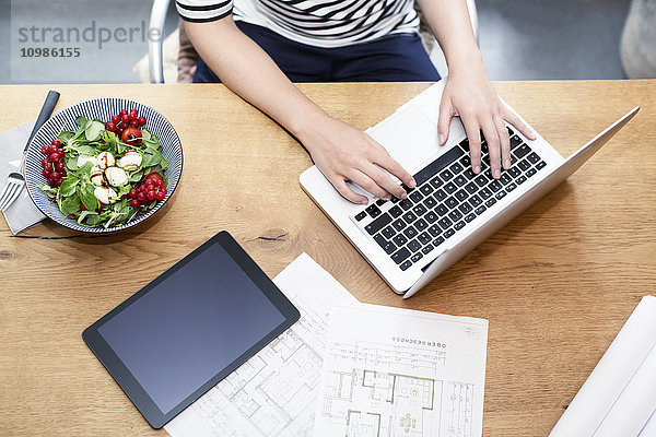Frau am Schreibtisch mit Laptop neben Bauplan und Salat