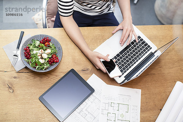 Frau am Schreibtisch mit Laptop und Handy neben Bauplan und Salat