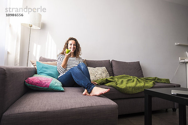 Porträt einer lächelnden Frau  die sich auf der Couch entspannt und einen Apfel isst.