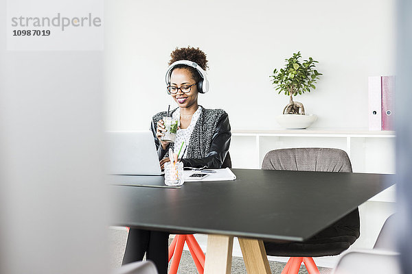 Geschäftsfrau mit Kopfhörer und Getränk am Schreibtisch sitzend mit Blick auf Laptop