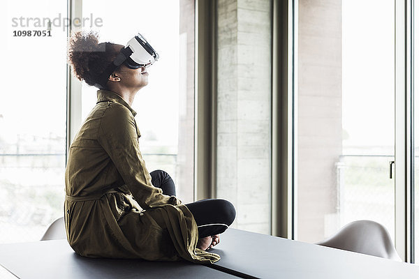 Frau im Büro sitzt auf einem Konferenztisch und trägt eine Virtual-Reality-Brille.