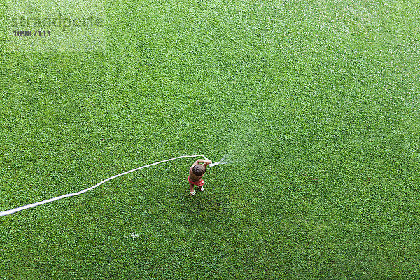 Kleiner Junge auf dem Rasen stehend mit Gartenschlauch  Draufsicht