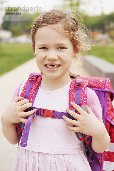 Porträt eines 5-jährigen Schulmädchens mit rosa und lila Schultasche  das lächelnd in die Kamera schaut und zeigt  dass ihr die Vorderzähne fehlen
