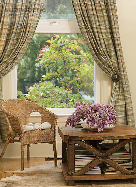 Stuhl und Couchtisch mit Blumen am Fenster mit Vorhängen im Wohnzimmer