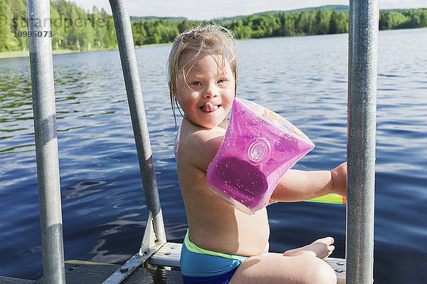 Mädchen mit Down-Syndrom trägt Schwimmflügel und sitzt auf dem Steg eines Sees  Schweden