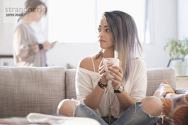 Junge Frau sitzt auf dem Sofa und hält eine Tasse  eine andere Frau benutzt ein Mobiltelefon