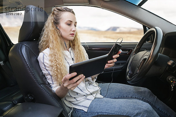 Frau sitzt im Auto und benutzt gleichzeitig Tablet und Mobiltelefon