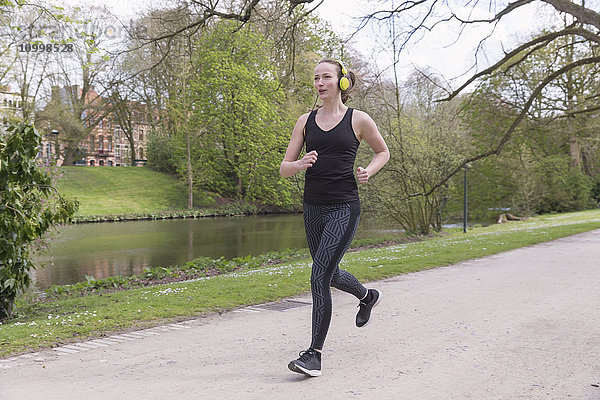 Mittelgroße Frau mit Kopfhörern joggt am Kanal im Park