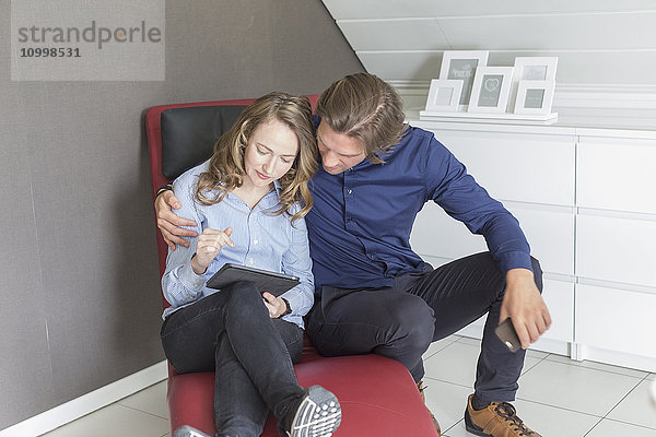 Paar im mittleren Erwachsenenalter sitzt auf einem Sessel und benutzt ein Tablet