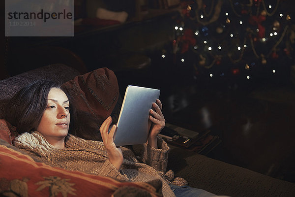 Frau mit digitalem Tablett entspannt auf dem Sofa im Wohnzimmer in der Nähe von Weihnachtsbaum