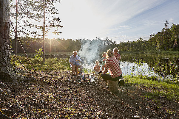 Großeltern und Enkelkinder beim Lagerfeuer am sonnigen Seeufer im Wald