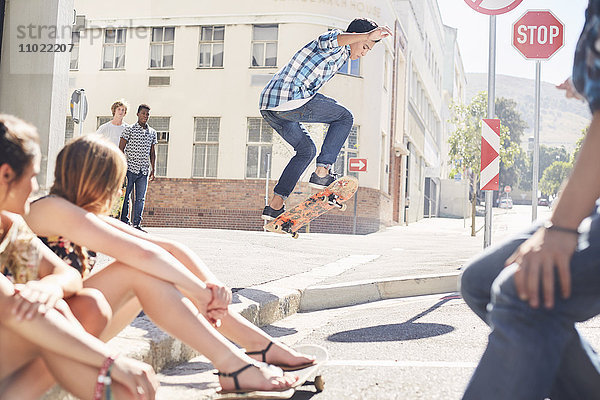 Freunde beobachten Teenager-Junge beim Skateboard-Springen an der sonnigen urbanen Ecke.