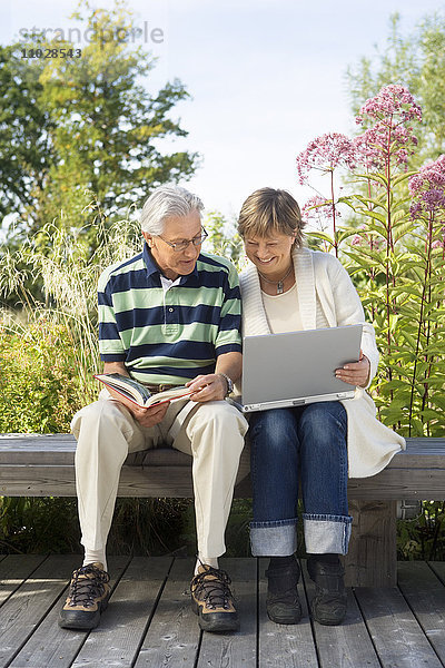 Eine Frau und ein Mann auf einer Bank im Freien mit einem Buch und einem Laptop.