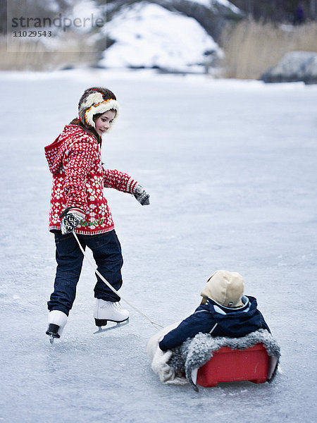 Mädchen  das seinen Bruder beim Schlittschuhlaufen auf einem zugefrorenen See auf einem Schlitten zieht