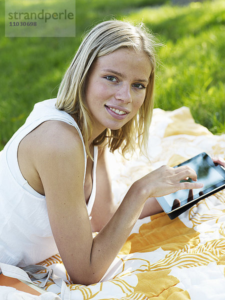 Junge Frau liegt auf einer Decke und hält ein digitales Tablet  lächelnd  Porträt