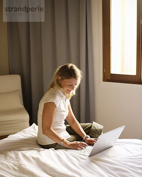 Skandinavische Frau sitzt auf dem Bett und benutzt einen Laptop.