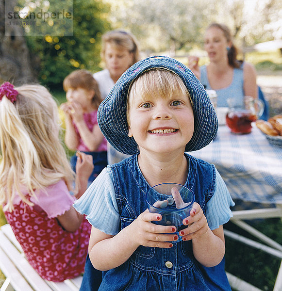 Ein lächelndes Mädchen mit Sonnenhut trinkt zusammen mit anderen Kindern und Erwachsenen Limonade.