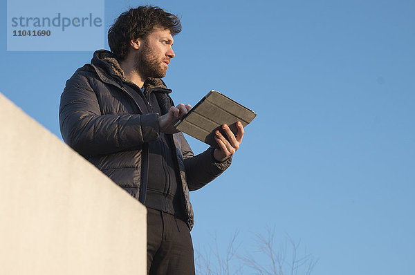 Mann hält digitales Tablett im Freien  schaut in Gedanken weg
