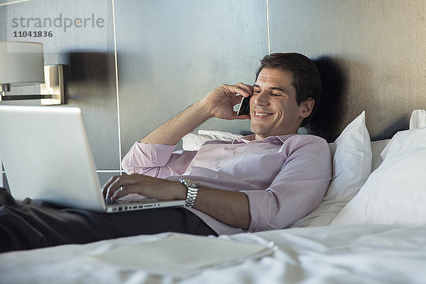 Mann im Bett liegend  mit Handy und Laptop-Computer